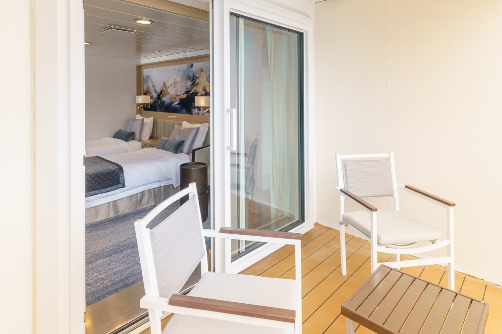View of balcony showing 2 chairs, small table and open sliding door with double bed inside of cabin.