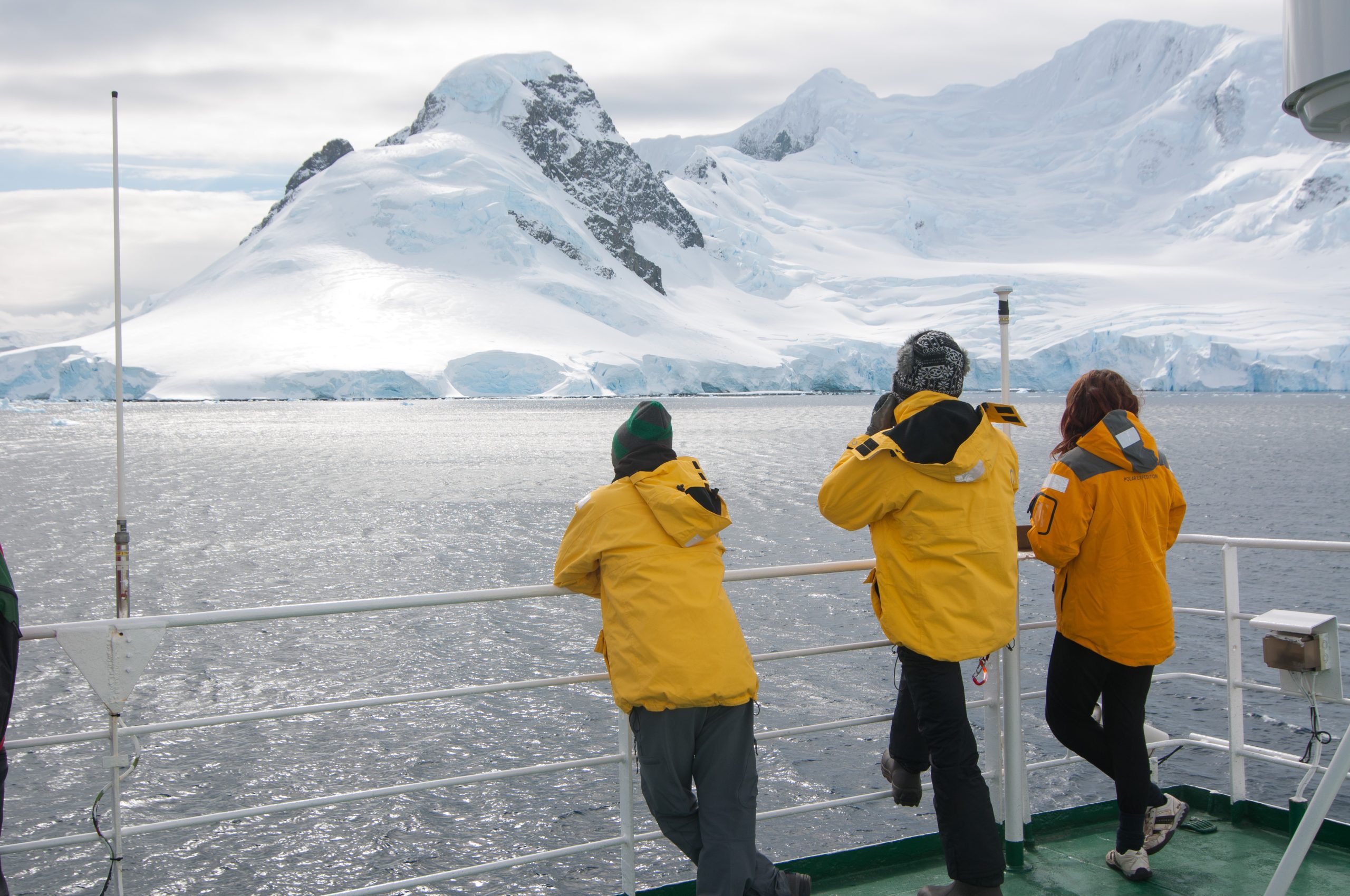 Three passengers standing on deck looking at snow covered mountain.