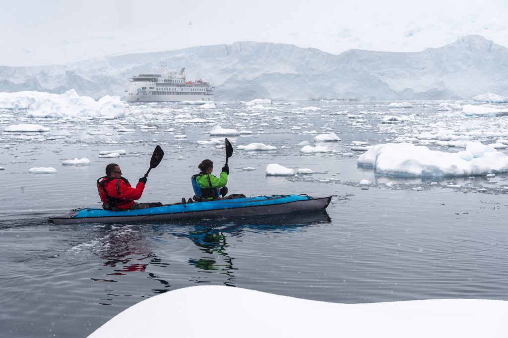 Two guest paddling in icy Antarctic waters with Greg Mortimer ship in the background