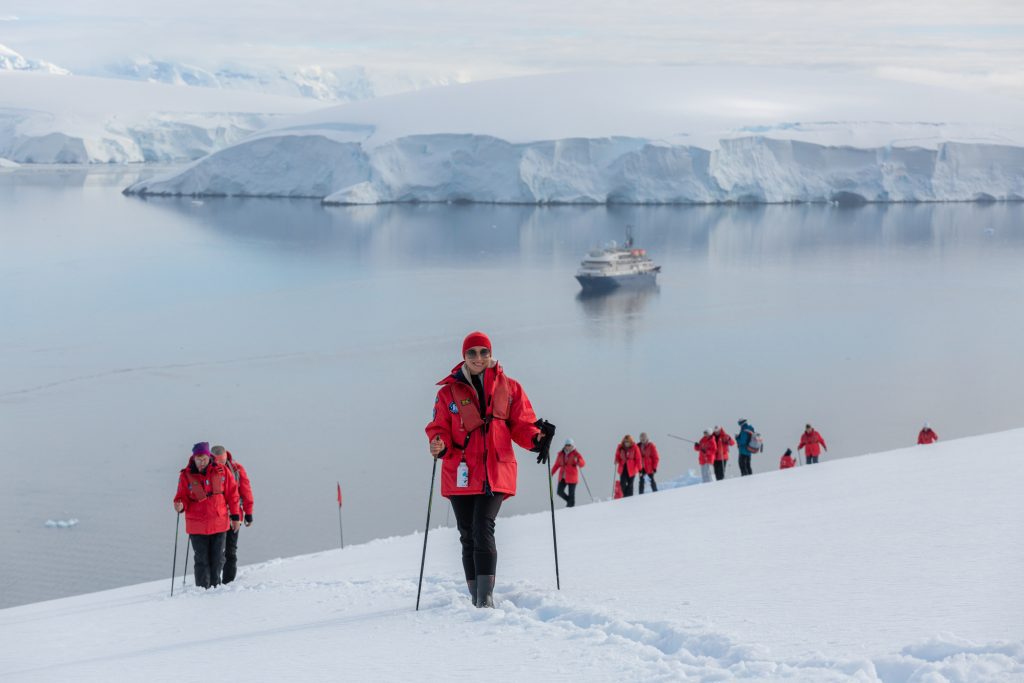Guests standing on steep snowy hillside with ship in the background.