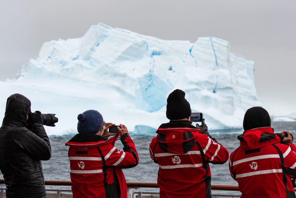 Guests standing on deck of ship taking photos of large iceberg.